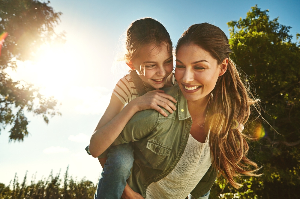 Mother piggy backing her daughter while smiling outside in the sunshine.